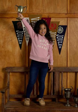 Naeratav tüdruk seisab puidust pingil, hoides hõbedast karikat pea kohale, taustal on puidust sein ja värvikad Joulesi lipud.