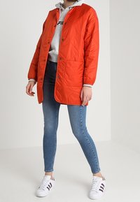 Pomarańczowa pikowana kurtka z zatrzaskami, noszona na szarej bluzie z kapturem. Połączona z niebieskimi dżinsami i białymi sneakersami z ciemnymi paskami.