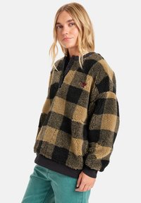 Fuzzy ruitenpatroon pulloverjack in tan en zwart met een kwartknopen sluiting, voorzien van een zachte textuur en geribde manchetten.