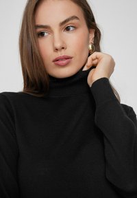 Suéter negro de cuello alto con textura de punto fino, mangas largas y silueta ajustada. Un modelo sostiene el cuello con una mano.