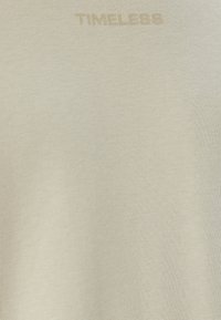 Beige katoenen T-shirt met een zachte textuur, voorzien van het woord "TIMELESS" in een lichte kleur gebosseleerd nabij de halslijn.