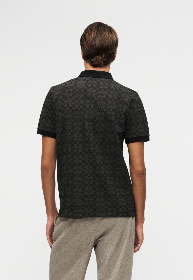 Man wearing black patterned polo shirt and beige pants, standing facing away against plain light background.