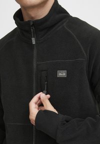 Schwarze Fleecejacke mit hohem Kragen, Reißverschluss und einer kleinen Brusttasche. Enthält strukturierten Stoff und gerippte Bündchen.