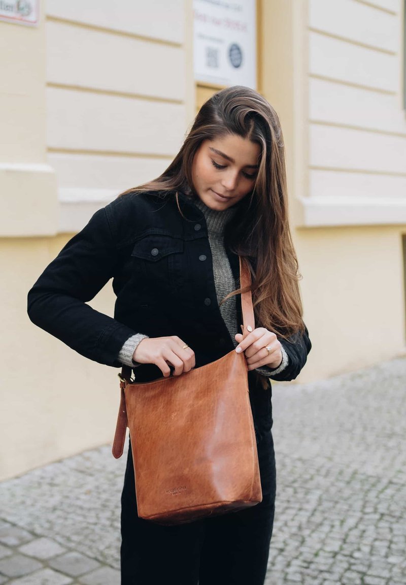 Braune Leder-Schultertasche mit glatter Textur und minimalistischem Design, ausgestattet mit einem einzigen Tragegurt und einem offenen Oberteil. Die Tasche ist leicht heruntergebeugt.