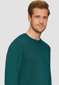 Donkergroene langemouwshirt met een ronde hals en een klein logo-patch op de borst, gemaakt van een zachte, gestructureerde stof.