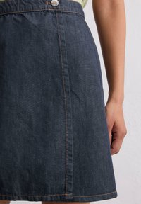 Jupe A-line en denim bleu foncé avec des coutures orange, dotée d'une fermeture à boutons à l'avant et d'un détail de couture latérale visible. Texture lisse.