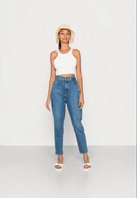 Top cropped branco de malha, jeans azuis de cintura alta e sandálias brancas. A modelo usa um chapéu de palha bege com detalhes em forma de flor amarela.