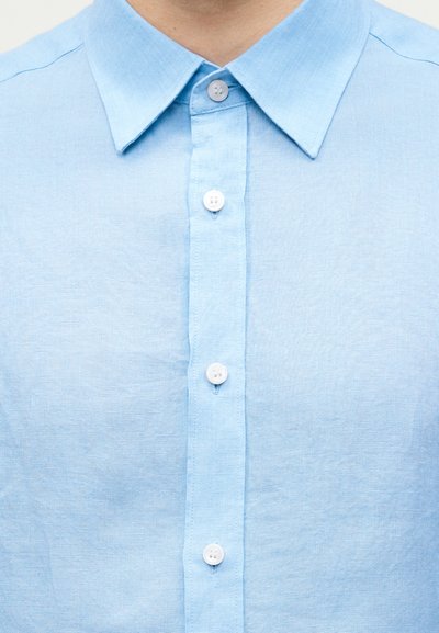 Ανοιχτό μπλε πουκάμισο με γιακά, με τέσσερα λευκά κουμπιά μπροστά, που φοριέται από ένα άτομο.