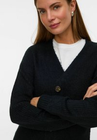 Tamna pletena kardigan jakna s V-izrezom, koja se zatvara jednim smešnim braon dugmetom i ima rebraste manžetne. Nosiva preko bele košulje.