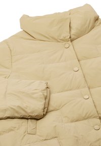 Lyse beige pufferjakke med høj krave, der har runde knapper, blød tekstur og diamant-quiltet mønster på stoffet.