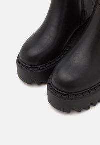 Gros plan sur des bottes en cuir noir avec des semelles plateformes épaisses et rainurées et des bouts arrondis sur un fond clair.