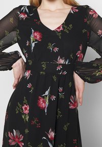 Robe noire à fleurs avec des motifs de fleurs rouges et bleues, manches longues transparentes, décolleté en V profond et taille cintrée.