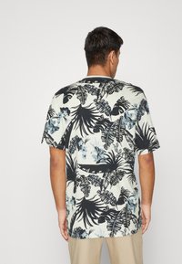 Krátký rukáv, světle zbarvená košile s černým a modrým tropickým listovým vzorem na texturované tkanině. Kulatý výstřih se rovnými lemy.