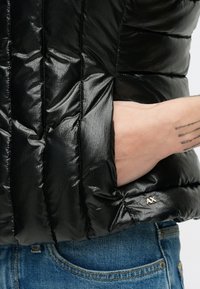 Čierna vatovaná bunda s lesklým povrchom, prešívaným dizajnom, bočným vreckom a zlatým logom "AX" pri leme. Nosená cez džínsové nohavice.