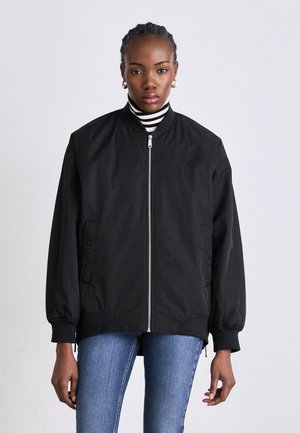 Giubbotto bomber nero con zip frontale, polsini e orlo a costine. Presenta tasche laterali e una texture di tessuto liscia e opaca. Sotto, un dolcevita a righe.