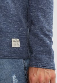 Marinblå texturerad långärmad skjorta med en liten vävd etikett vid fållen, tillsammans med ljusa denimjeans som visar ett slitet mönster.