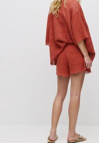 Kaftan ample couleur rouille associé à un short assorti. Tissu léger et texturé avec des bords en forme de coquille et des détails subtils.