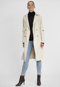 Beige színű trench coat, dupla gombolással, széles hajtókákkal és felgöngyölt ujjakkal, kék skinny farmerral és sötét bokacsizmával párosítva.
