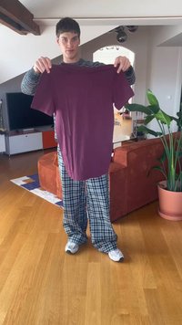 Jaunais cilvēks tur rokās purpura krāsas T-kreklu izstieptu priekšā, stāvot viesistabā ar koka grīdu un laukuma raksta pidžamu biksēm.