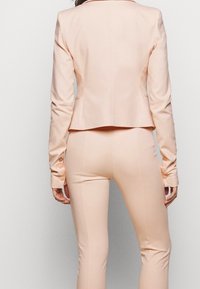 Bladoróżowa dopasowana marynarka z usztywnionym kołnierzem, wąskimi rękawami zdobionymi guzikami, w zestawie z dopasowanymi, szytymi na miarę spodniami. Gładka faktura.