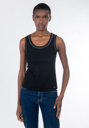 Donna che indossa un top nero senza maniche con borchie d'argento lungo lo scollo, abbinato a jeans blu, in piedi davanti a uno sfondo bianco.