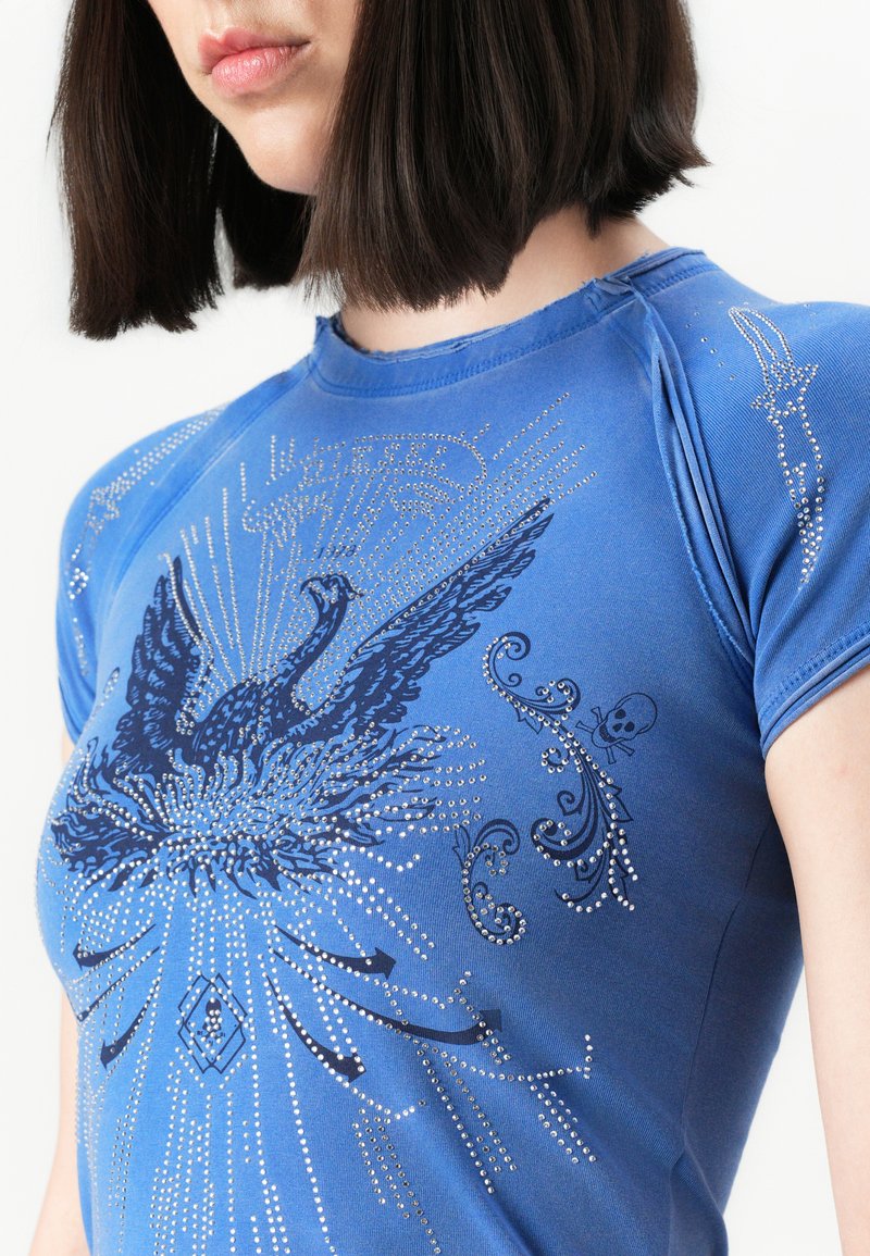 Woman wearing blue T-shirt with black stylized bird and skull designs decorated with small silver studs.