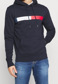 Osoba v námořnickém modrém mikině s červeným, bílým a modrým logem Tommy Hilfiger přes hruď a modrých džínech.