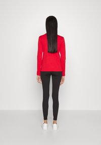 Röd långärmad topp i kombination med svarta leggings. Slät textur, åtsittande design. Vita sneakers avslutar outfiten. Bakifrån vy visas.