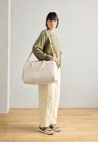 White leather duffle bag with dual handles and adjustable strap, featuring a smooth texture and subtle stitching details. Model stands against a light background.