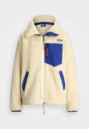 Veste en polaire couleur crème avec poche poitrine zippée bleue et poches latérales, fermeture éclair devant et col montant.