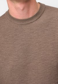 Maglione marrone confezionato a maglia con una superficie strutturata, scollatura rotonda a coste e un design aderente. Nessun logo o motivo visibili.