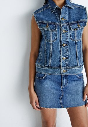 Person iført blå denim vest uden ærmer og matchende denim nederdel med frynset kant, stående mod hvid baggrund.