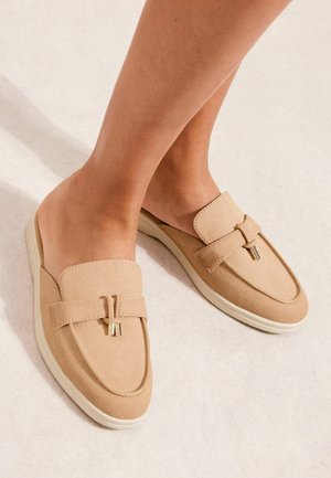 Beżowe wsuwane loafersy z ozdobnym paskiem i metalicznymi frędzlami, noszone na bose stopy na jasnym tle.