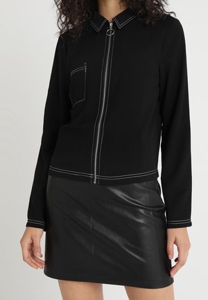 Kvinde iført sort lynlåstrøje med hvide syninger og sort lædermininederdel, der står mod en ensfarvet baggrund.