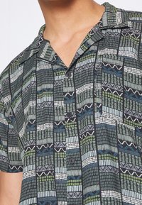 Camisa de manga corta con un patrón geométrico en verde, negro, blanco y azul; con botones en la parte delantera y un diseño de cuello relajado.