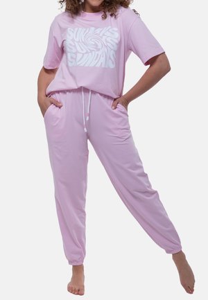 Persoon draagt een roze loungewear-set met “SUNDAYS” drie keer gedrukt op het shirt, handen in de zakken, staat op één been op blote voeten.