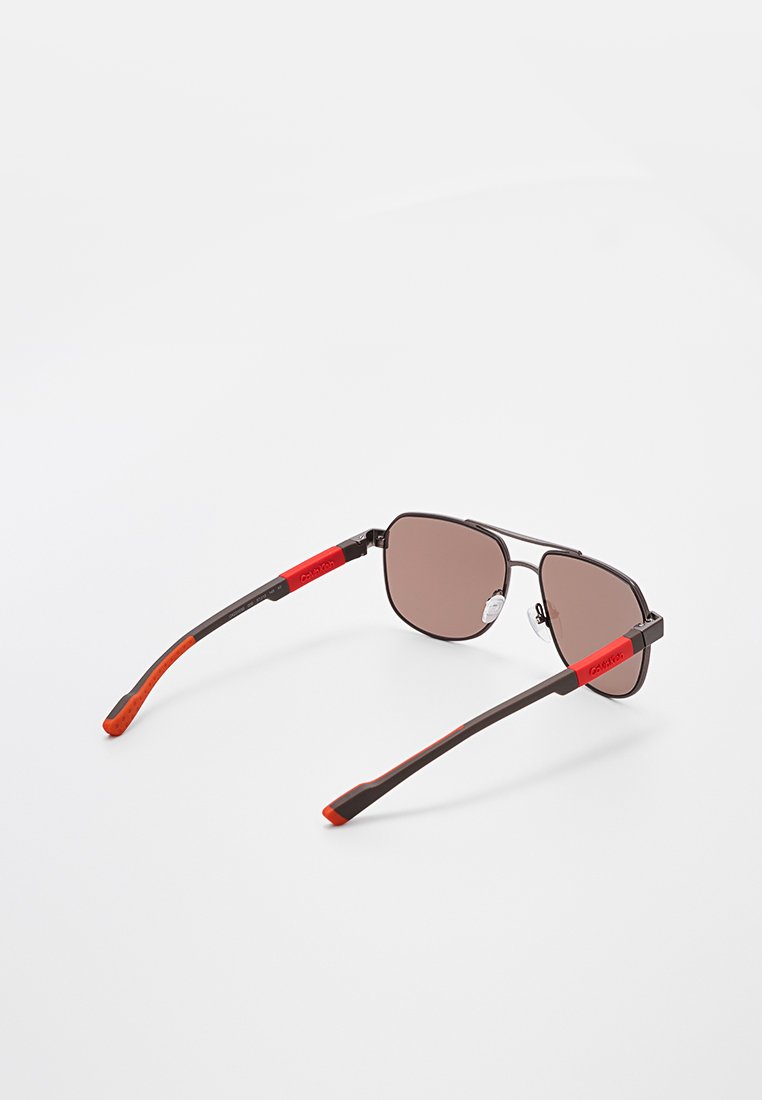 Gafas de sol de marco negro elegante con lentes marrones y detalles rojos en las varillas, colocadas sobre una superficie blanca en un ángulo que muestra la parte trasera.