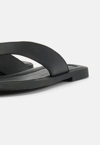 Κοντινό πλάνο μαύρου επίπεδου σανδαλιού με φαρδιά δερμάτινη λωρίδα και μαλακή σόλα σε λευκό φόντο.