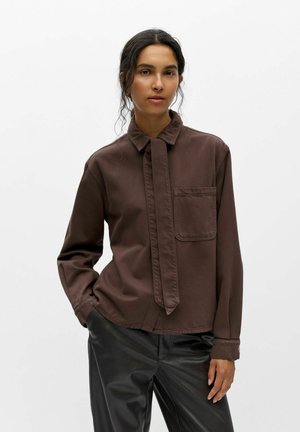Femme aux cheveux foncés portant une chemise marron à boutons avec une poche avant et un pantalon en cuir noir sur un fond blanc uni.