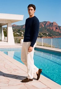 Homme portant un pull bleu marine, un pantalon blanc et des chaussures marron, debout près d'une piscine avec une rambarde, des montagnes et un ciel dégagé en arrière-plan.