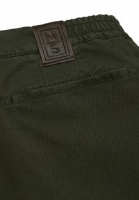 Donkergroene broek met een gestructureerde stof, elastische tailleband en een bruine leren label met 'M5' opgedrukt. Achterzak zichtbaar.