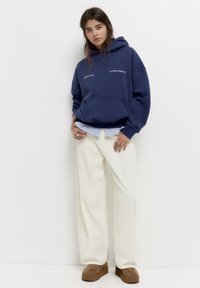 Marineblauwe hoodie met een voorzak en witte tekst, gelaagd over een lichtblauw gestreept shirt. Gecombineerd met crèmekleurige wijde broek en bruine plateau-schoenen.