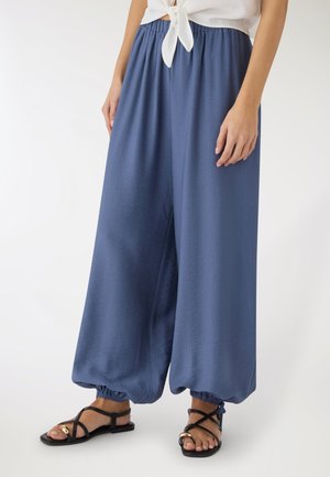 Donna che indossa pantaloni blu larghi con caviglia elastica, camicia bianca annodata e sandali neri bassi con accenti dorati, in piedi su uno sfondo bianco.