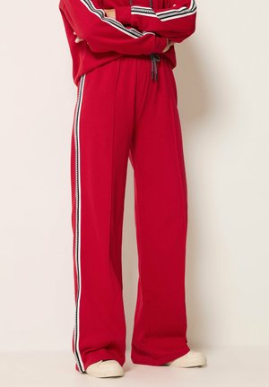 Persona con pantalones deportivos rojos con franjas laterales blancas y negras y zapatillas blancas, de pie con los brazos cruzados.