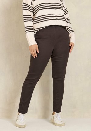 Persona con pantalones ajustados de color marrón oscuro, zapatillas blancas y un suéter blanco con rayas horizontales negras, de pie frente a un fondo beige.