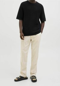 Černý texturovaný top s krátkým rukávem, béžové zúžené kalhoty, černé sandály s pásky, minimalistický design, na neutrálním pozadí.