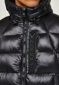 Parka nera con colletto alto, caratterizzata da una texture trapuntata, una zip frontale e un dettaglio in nastro verticale con marchio sul petto.