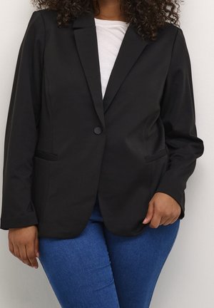 Kvinde iført en sort blazer med en enkelt knap over en hvid top og blå jeans, stående mod en ensfarvet lys baggrund.