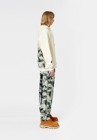 Sudadera beige con un patrón texturizado en verde y gris en la parte posterior; combinada con pantalones de camuflaje, que tienen rayas laterales y zapatos color beige.