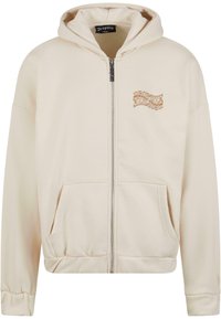 Cream-colored zip-up hoodie features a kangaroo pocket, ribbed cuffs, and an embroidered logo in brown on the left chest. Soft cotton material.